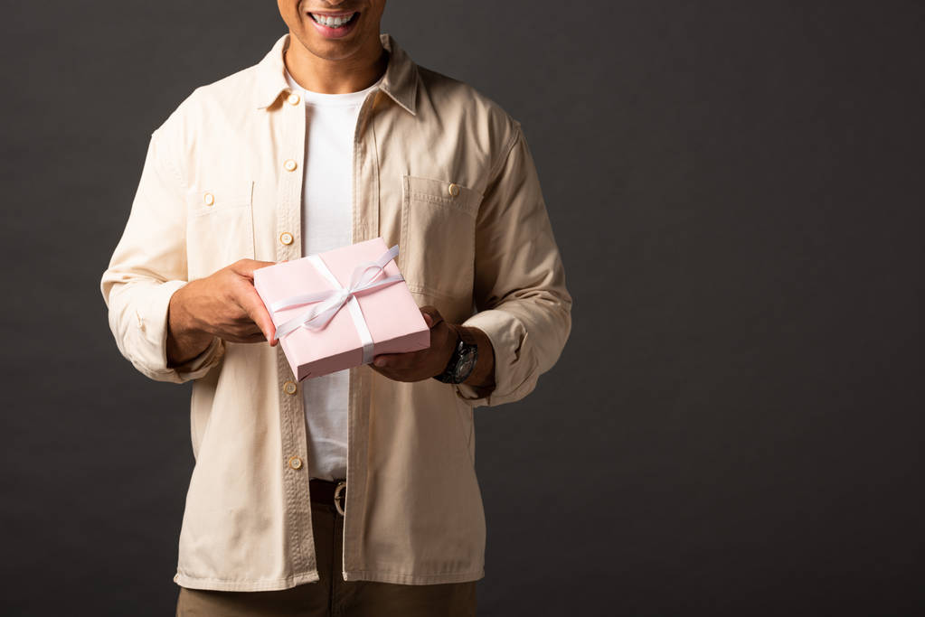 abgeschnittene Ansicht eines lächelnden Mischlingsmenschen im beigen Hemd mit Geschenk auf schwarzem Hintergrund - Foto, Bild