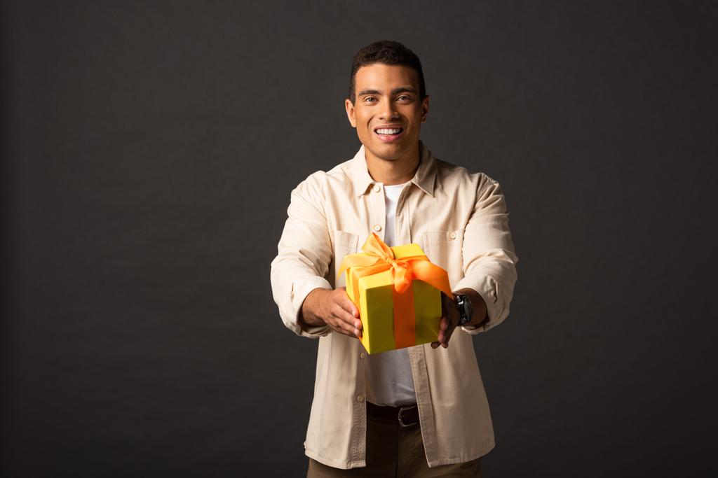 glimlachend knap gemengd race man in beige shirt feest cadeaus aanwezig op zwarte achtergrond - Foto, afbeelding