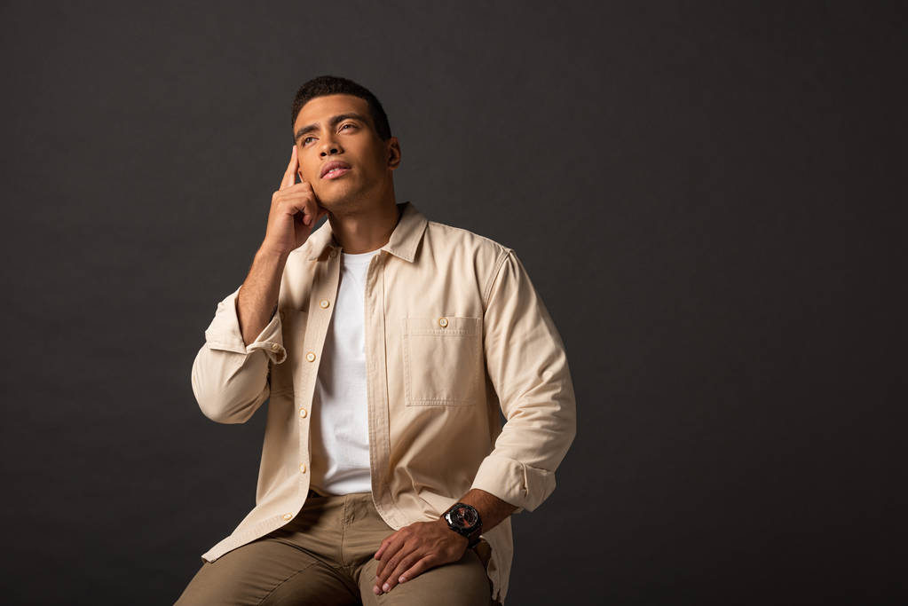 pensive handsome mixed race man in beige shirt sitting on chair on black background - Photo, Image