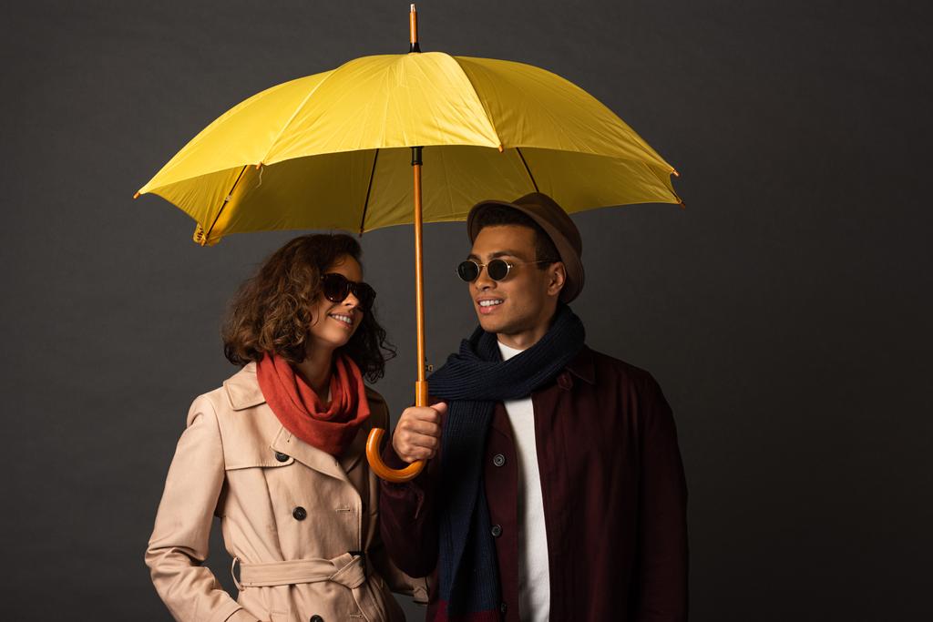 усміхнена стильна міжрасова пара в осінньому вбранні, що тримає жовту парасольку на чорному тлі
 - Фото, зображення