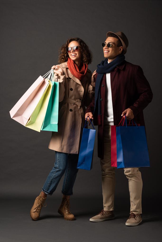 sonriente elegante pareja interracial en otoño traje sosteniendo bolsas de compras sobre fondo negro
 - Foto, imagen