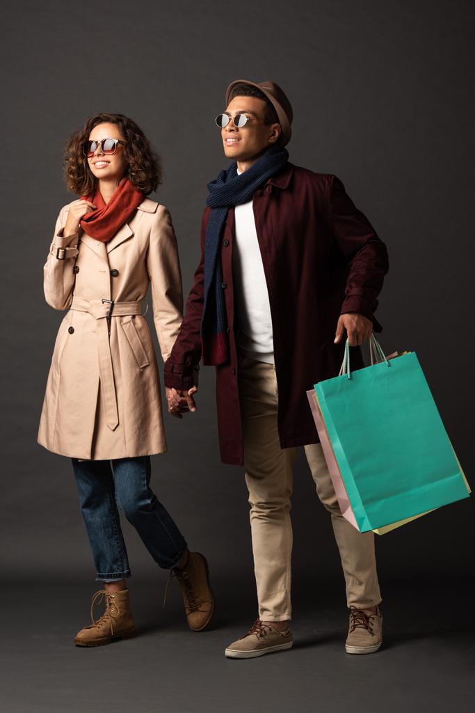 lächelndes stilvolles gemischtrassiges Paar im Herbst-Outfit mit Händchen und Einkaufstaschen auf schwarzem Hintergrund - Foto, Bild