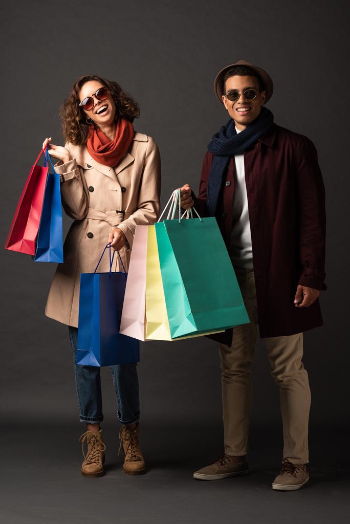 happy stylish interracial couple in autumn outfit holding shopping bags on black background - Photo, Image