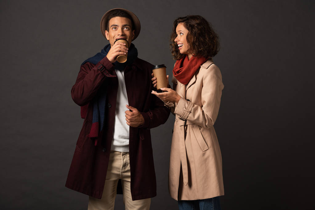 Улыбающаяся стильная межрасовая пара в осеннем наряде держит кофе, чтобы пойти и поговорить на черном фоне
 - Фото, изображение