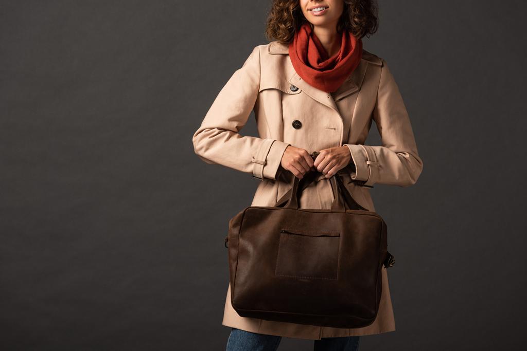 przycięty widok stylowej kobiety w płaszczu i szalik trzymając skórzaną brązową torbę na czarnym tle - Zdjęcie, obraz