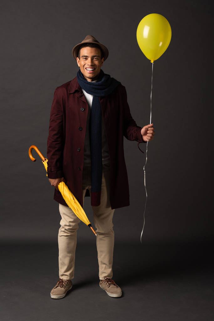 smiling mixed race man in scarf and hat holding yellow balloon and umbrella on black background - Photo, Image
