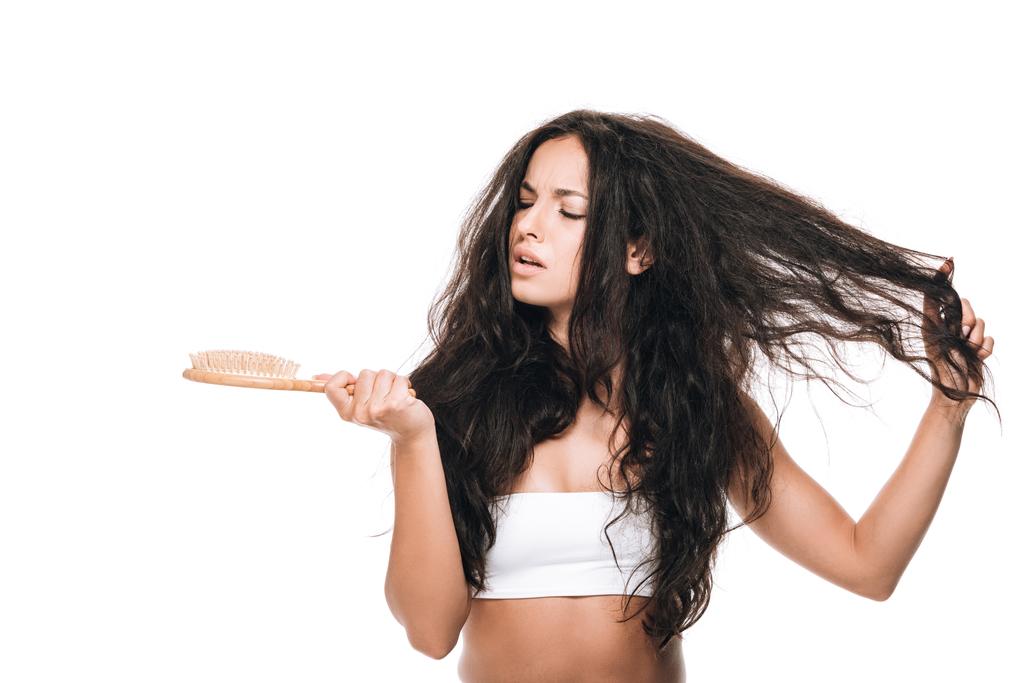 расстроенная брюнетка красивая женщина с волнистыми непослушными волосами глядя на расческу изолированы на белом
 - Фото, изображение