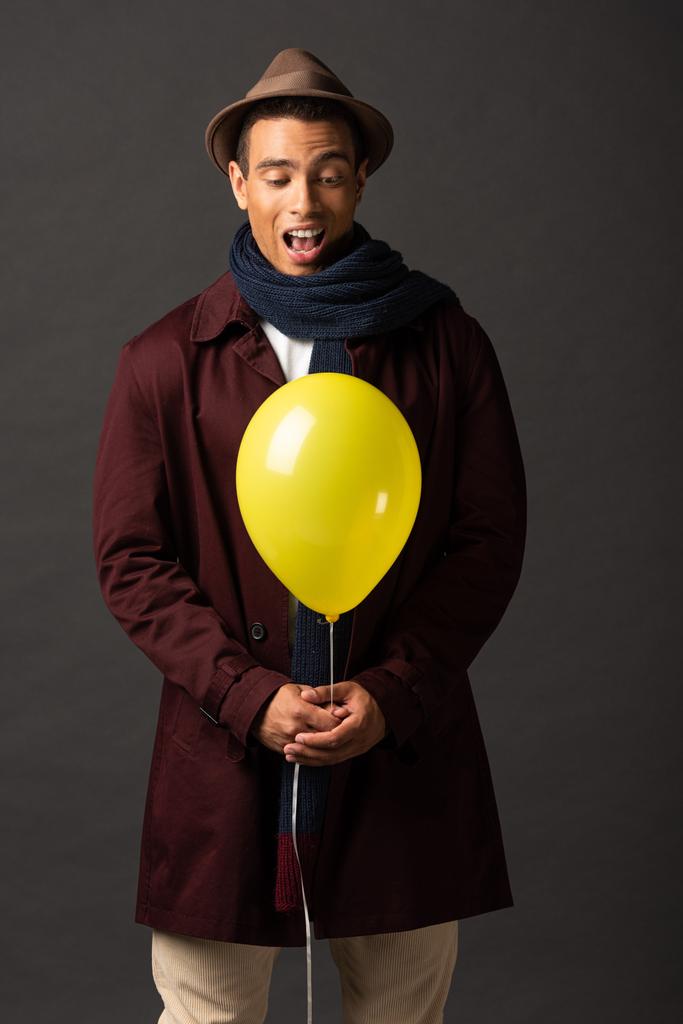 excited mixed race man in scarf and hat holding yellow balloon on black background - Photo, Image