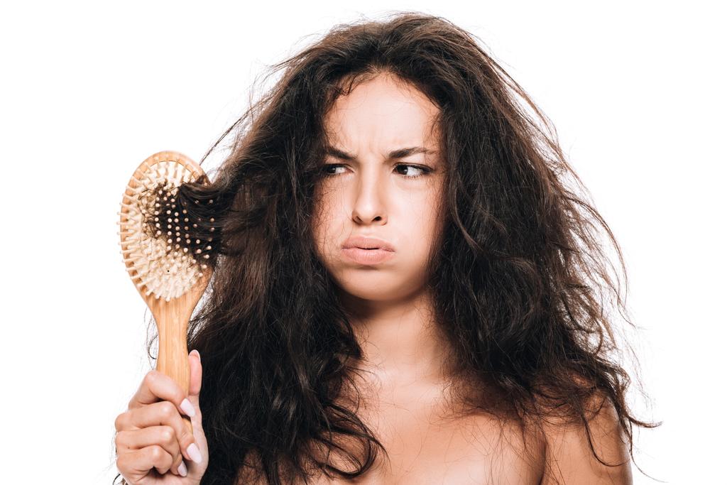 στρεσαρισμένη μελαχρινή γυναίκα κοιτάζοντας κυματιστό ατίθασο μαλλιά σε χτένα απομονωμένη σε λευκό - Φωτογραφία, εικόνα