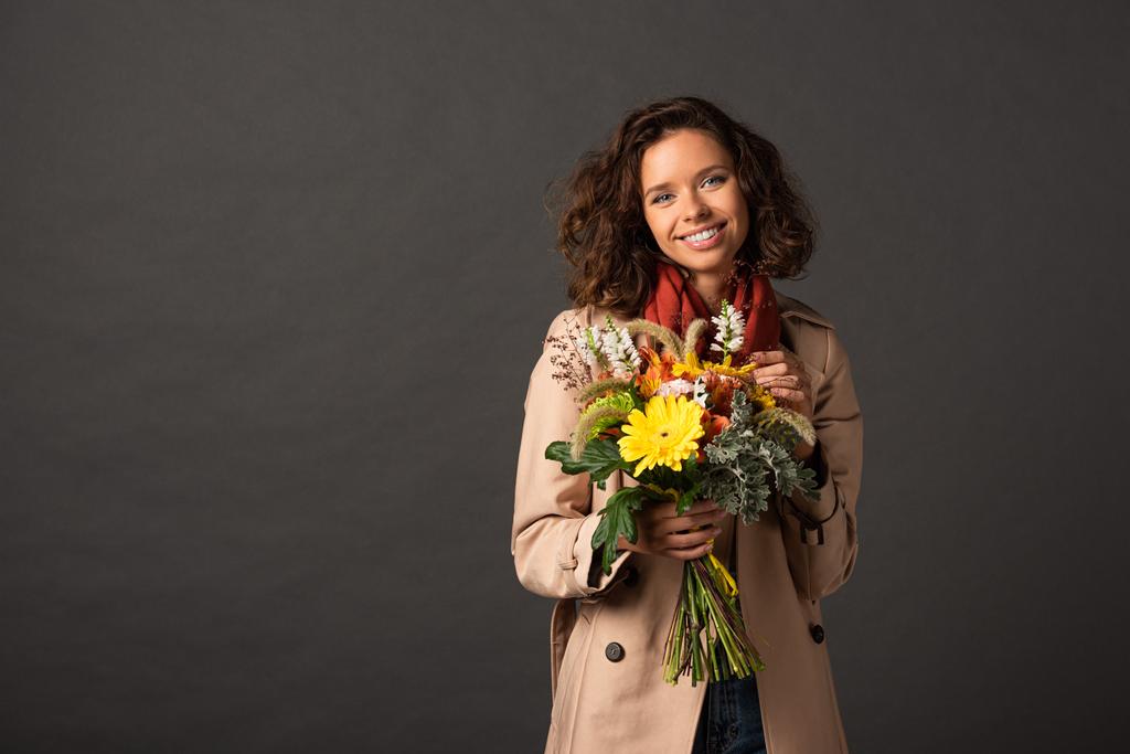 smiling curly woman in trench coat holding bouquet of autumnal wildflowers on black background - Photo, Image
