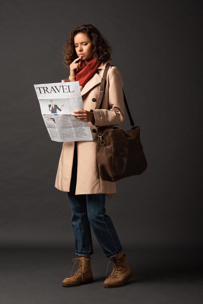 Nachdenkliche Frau im Trenchcoat mit Ledertasche liest Reisezeitung auf schwarzem Hintergrund - Foto, Bild