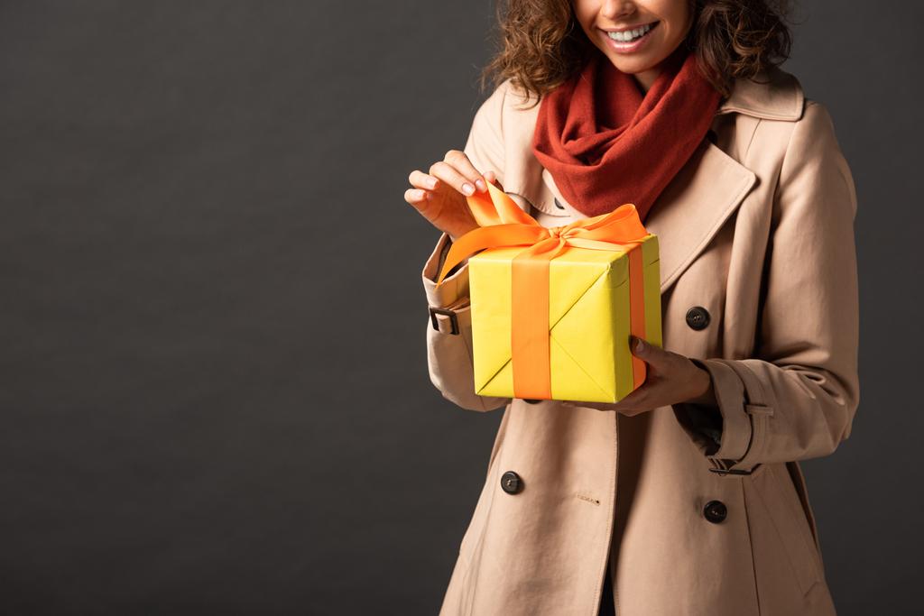 黒い背景にプレゼントを保持するトレンチコートで笑顔の女性のトリミングされたビュー - 写真・画像