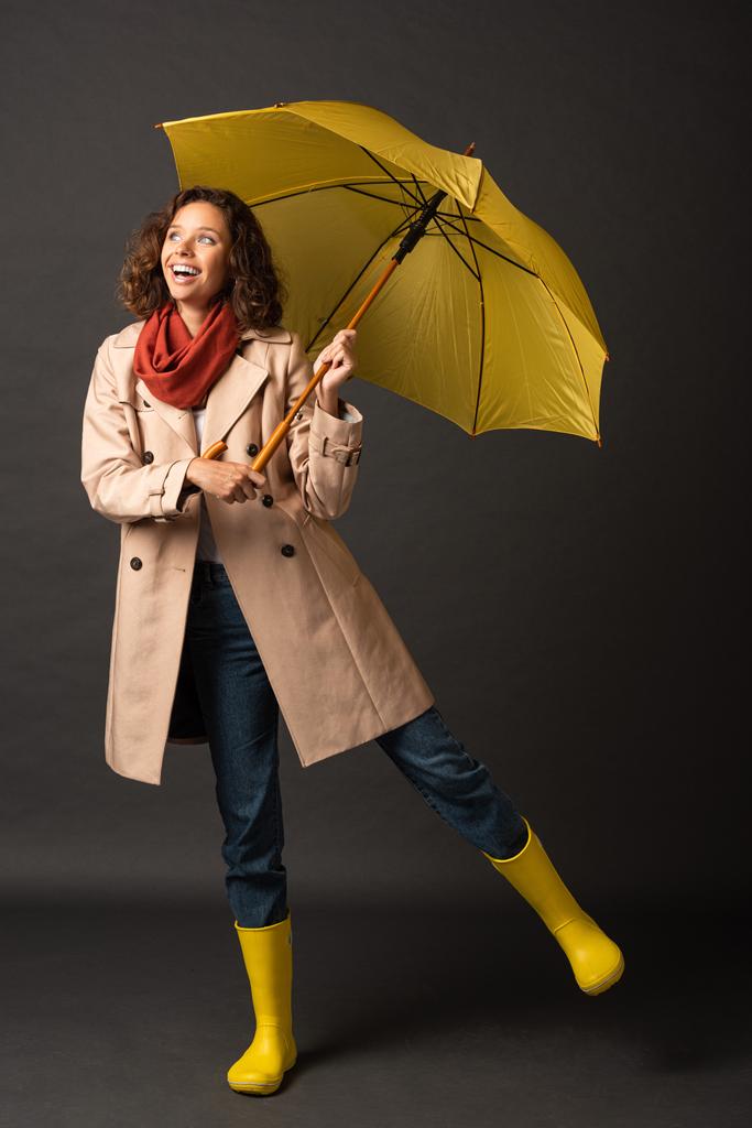 Gelukkige vrouw in trenchcoat en rubberen laarzen poseren met gele paraplu op zwarte achtergrond - Foto, afbeelding