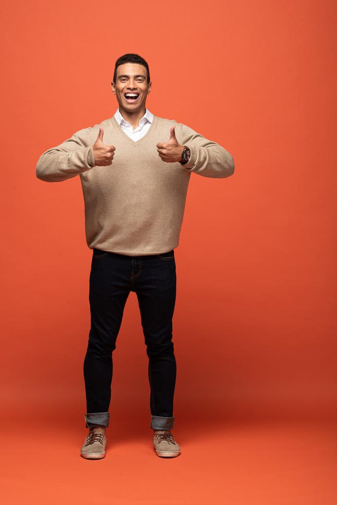 счастливый смешанный человек в бежевом свитере, показывающий большие пальцы на оранжевом
 - Фото, изображение