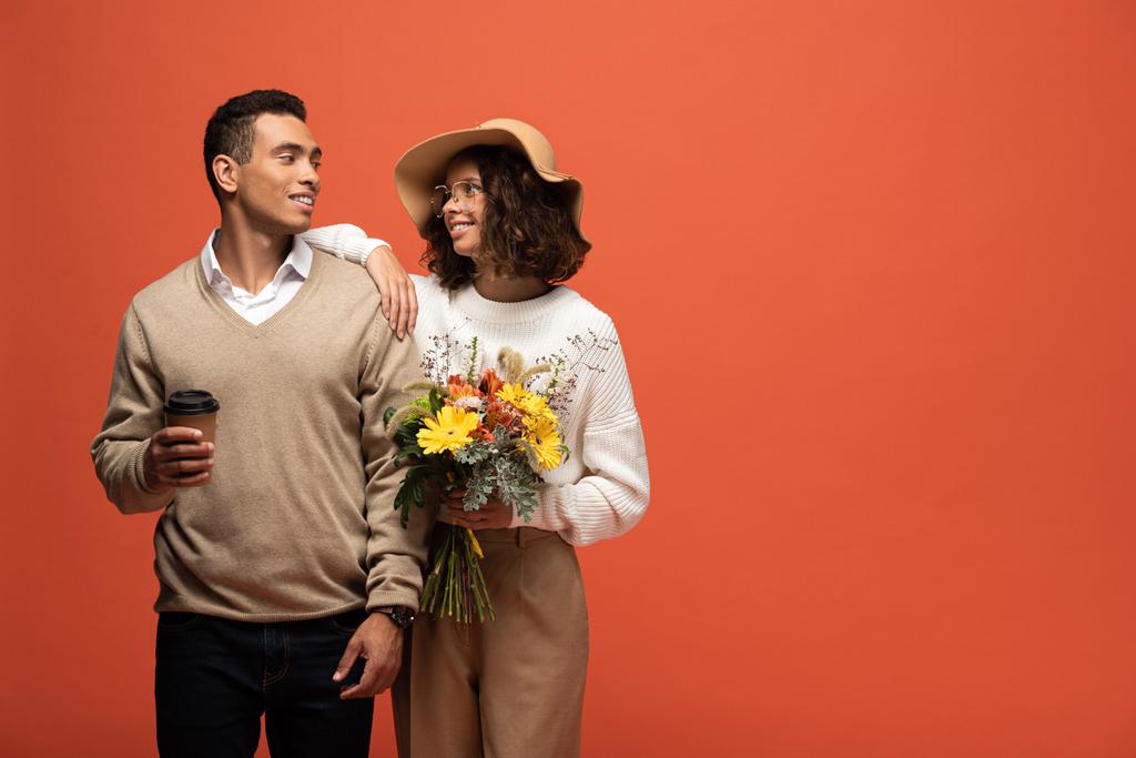 elegante casal interracial no outono roupa com café para ir e buquê de flores isoladas em laranja
 - Foto, Imagem