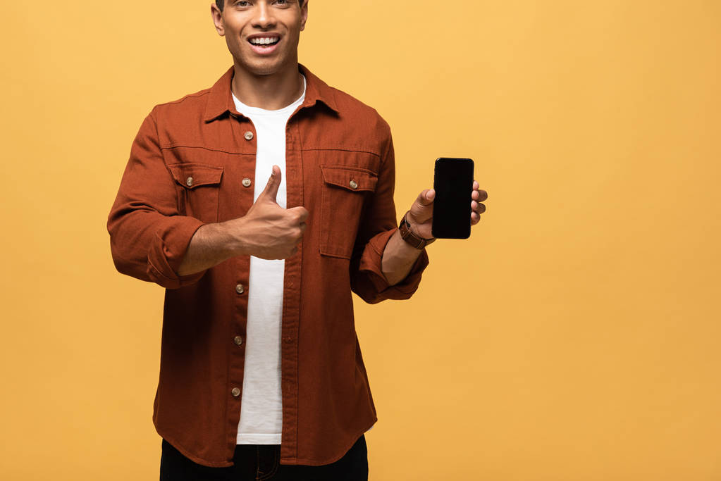 bijgesneden weergave van glimlachende mixed race man met smartphone met leeg scherm en weergeven duim omhoog geïsoleerd op geel - Foto, afbeelding