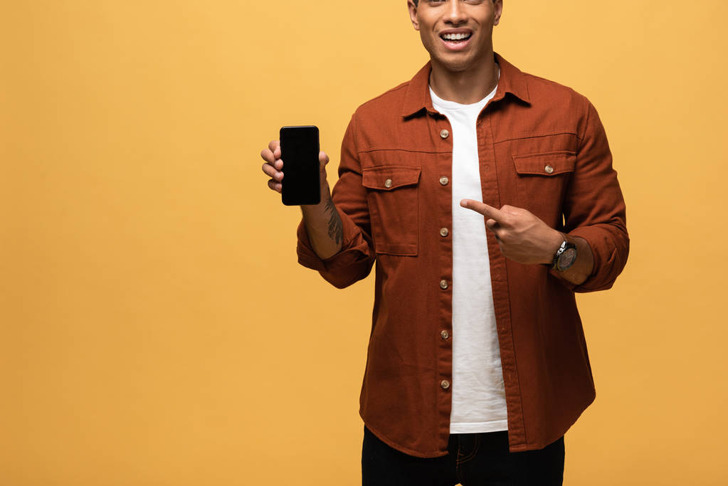 vista cortada do homem de raça mista apontando com o dedo para o smartphone com tela em branco isolada no amarelo
 - Foto, Imagem