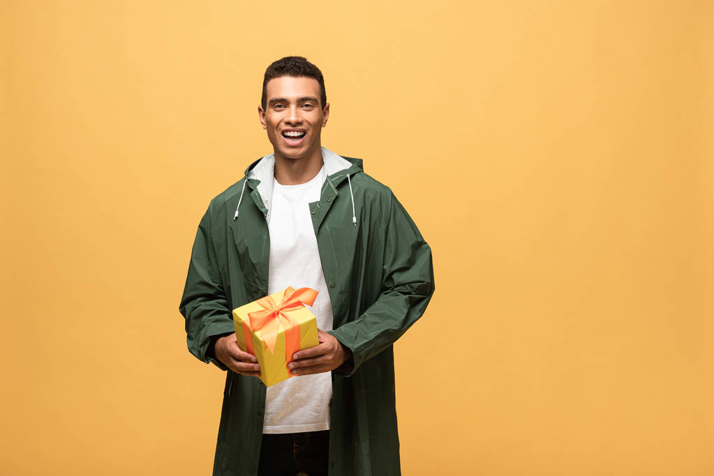 hombre de raza mixta feliz en impermeable y botas de goma que sostienen la caja de regalo aislada en amarillo
 - Foto, imagen