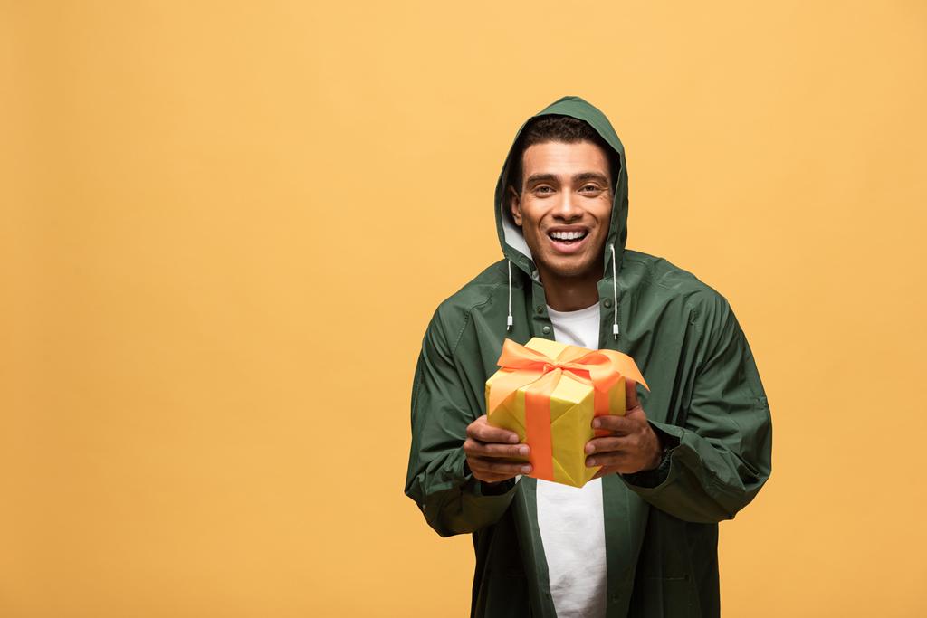 smiling mixed race man in raincoat and rubber boots holding gift box isolated on yellow - Photo, Image