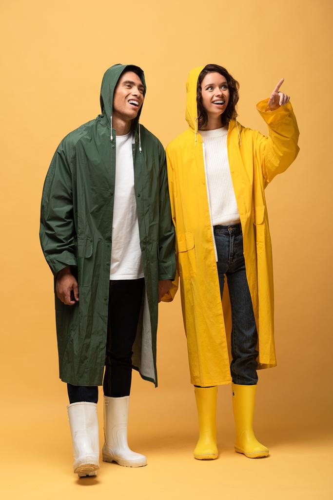 Gelukkig interracial paar in regenjassen en rubberen laarzen wijzend met vinger op gele achtergrond - Foto, afbeelding