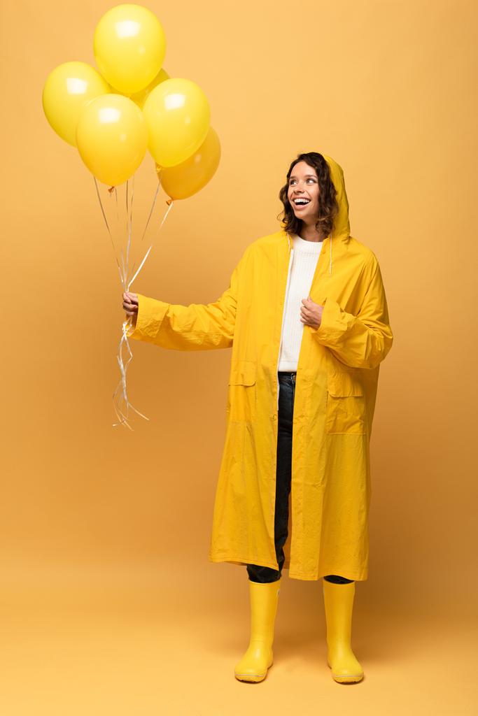 glückliche lockige Frau in gelbem Regenmantel und Gummistiefeln mit Luftballons auf gelbem Hintergrund - Foto, Bild