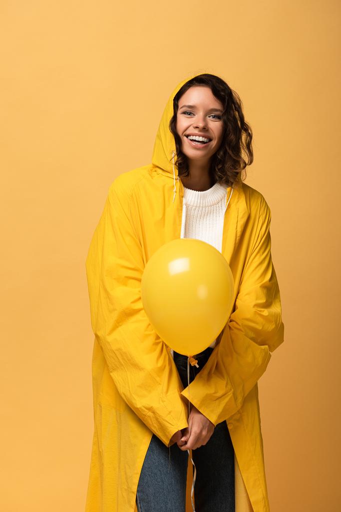 glückliche lockige Frau in gelbem Regenmantel mit Luftballon auf gelbem Grund - Foto, Bild
