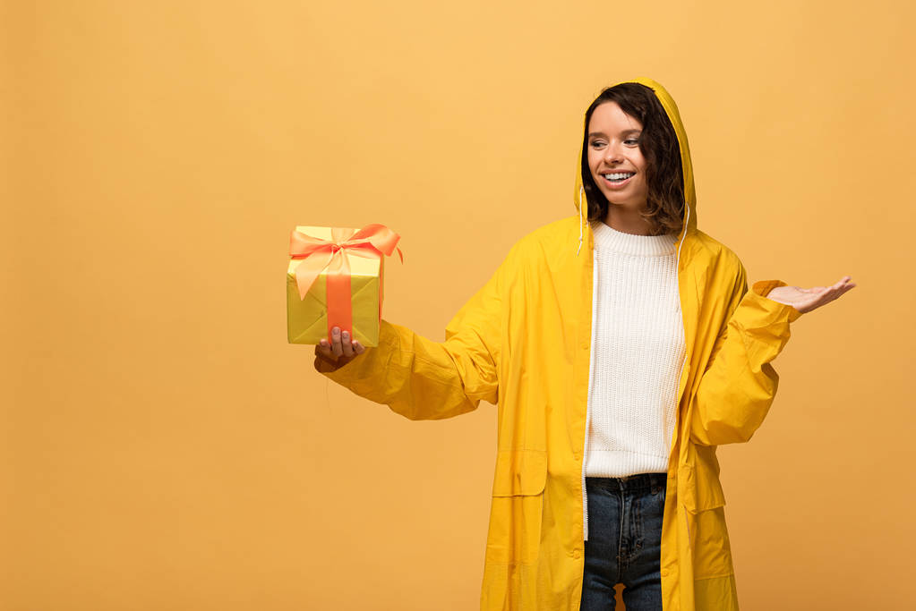 χαμογελαστή σγουρά γυναίκα σε κίτρινο αδιάβροχο κρατώντας κουτί δώρου απομονωμένο σε κίτρινο - Φωτογραφία, εικόνα