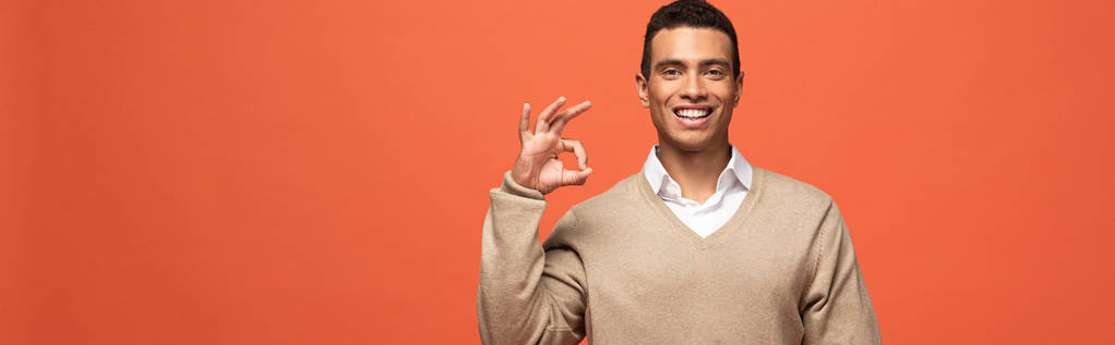 panoramic shot of happy mixed race man in beige sweater showing ok sign isolated on orange - Photo, Image