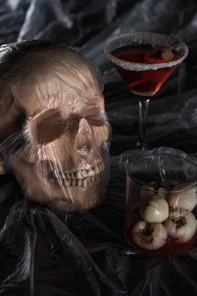 spooky human skull in cellophane near red cocktail on black background, Halloween decoration - Photo, Image