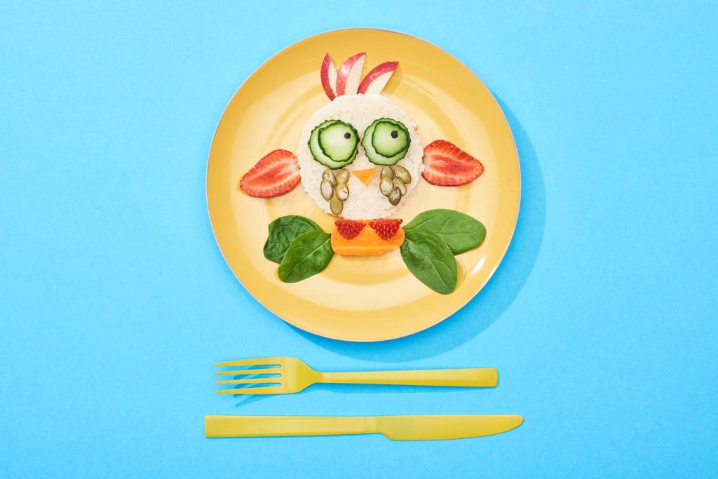 pohled na talíř s ozdobnou tváří, která je vyrobena z jídla pro děti v blízkosti příborů na modrém pozadí - Fotografie, Obrázek
