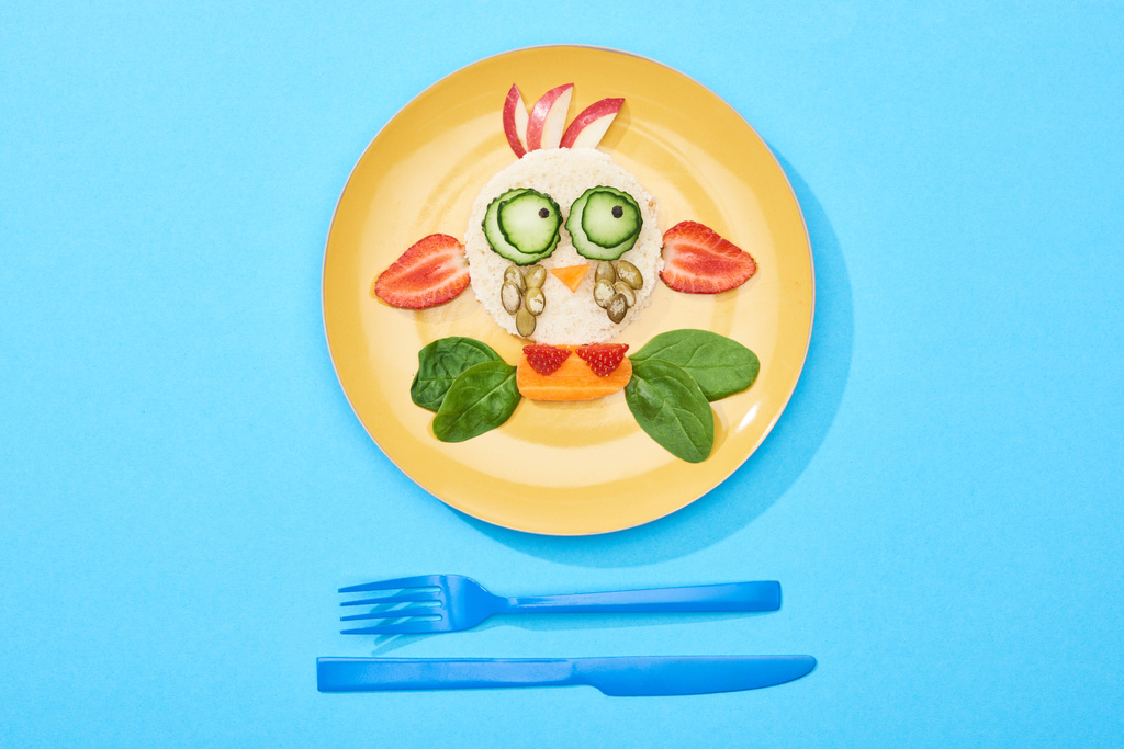 Teller mit ausgefallenem Gesicht aus Kinderfrühstück in der Nähe von Besteck auf blauem Hintergrund - Foto, Bild