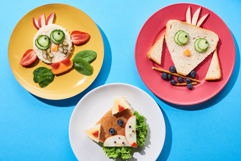 top view of plates with fancy cow, fox and bird made of food for childrens breakfast on blue background - Photo, Image