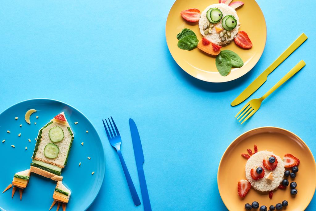top view of plates with fancy animals and rocket made of food for childrens breakfast near cutlery on blue background - Photo, Image