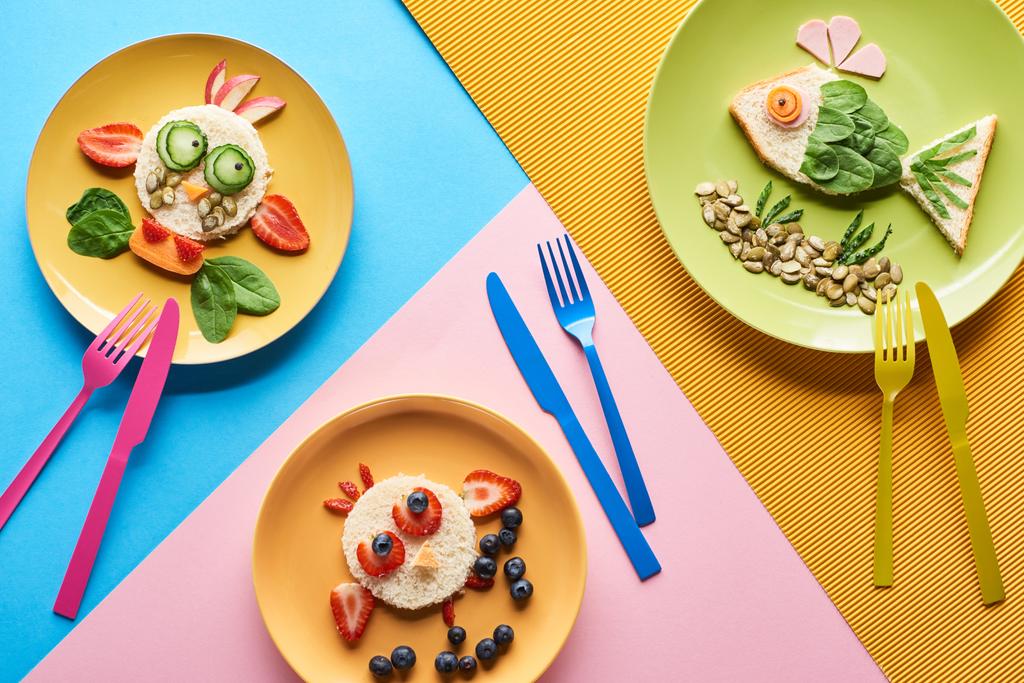 вид зверху на тарілки з вишуканими тваринами з їжі для дитячого сніданку на синьому, жовтому та рожевому тлі з столовими приборами
 - Фото, зображення