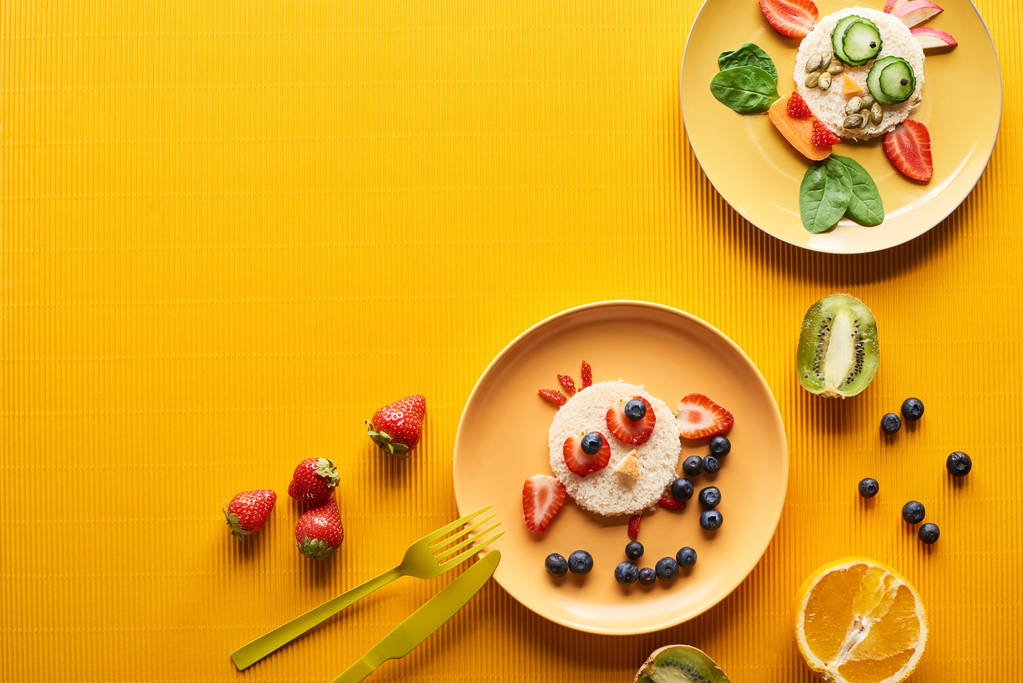 pohled na talíře s fantastovými zvířaty z potravin blízko příborů na barevném oranžovém pozadí - Fotografie, Obrázek
