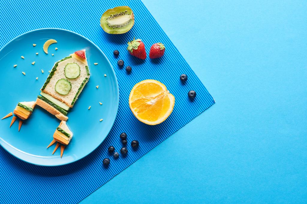 Teller mit ausgefallener Rakete aus Lebensmitteln auf blauem Hintergrund mit Früchten von oben - Foto, Bild