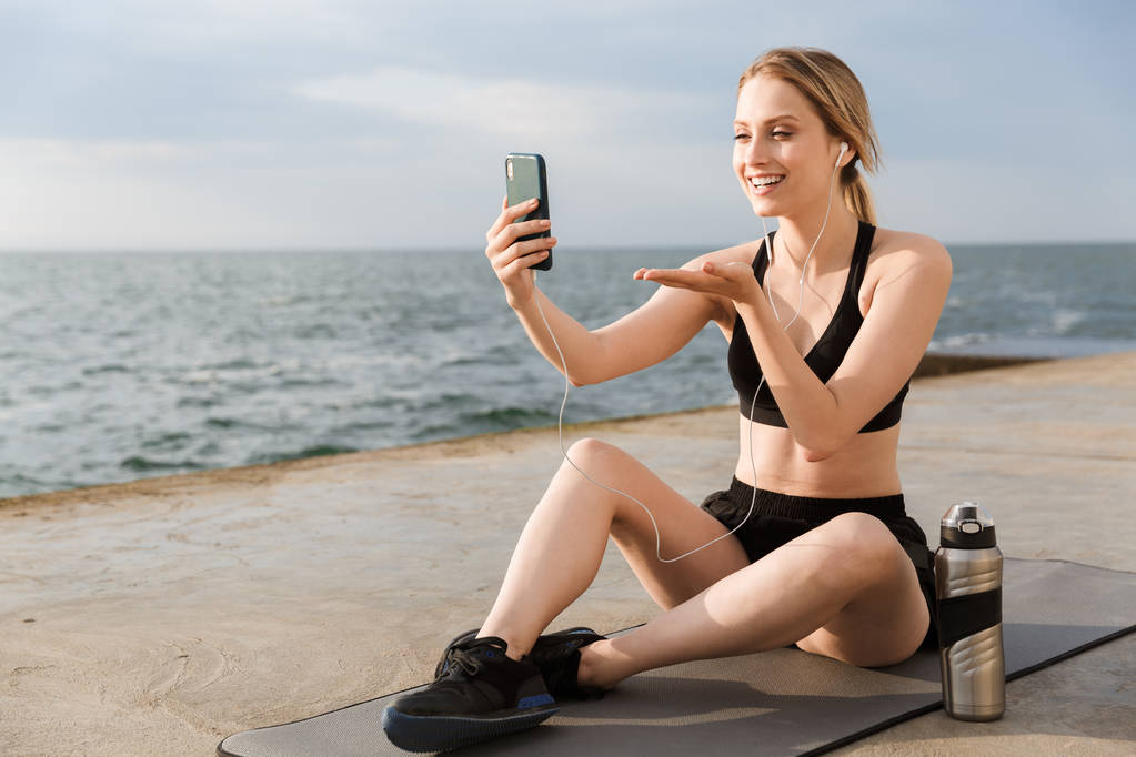 Imagen de la mujer alegre tomando selfie en el teléfono celular y sosteniendo el espacio de copia mientras se sienta en la estera cerca de la playa
 - Foto, imagen