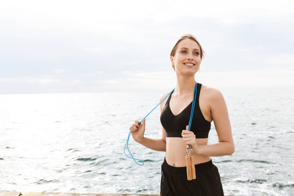 Afbeelding van lachende vrouw met springtouw over de nek tijdens het trainen in de buurt van Seaside in Morning - Foto, afbeelding