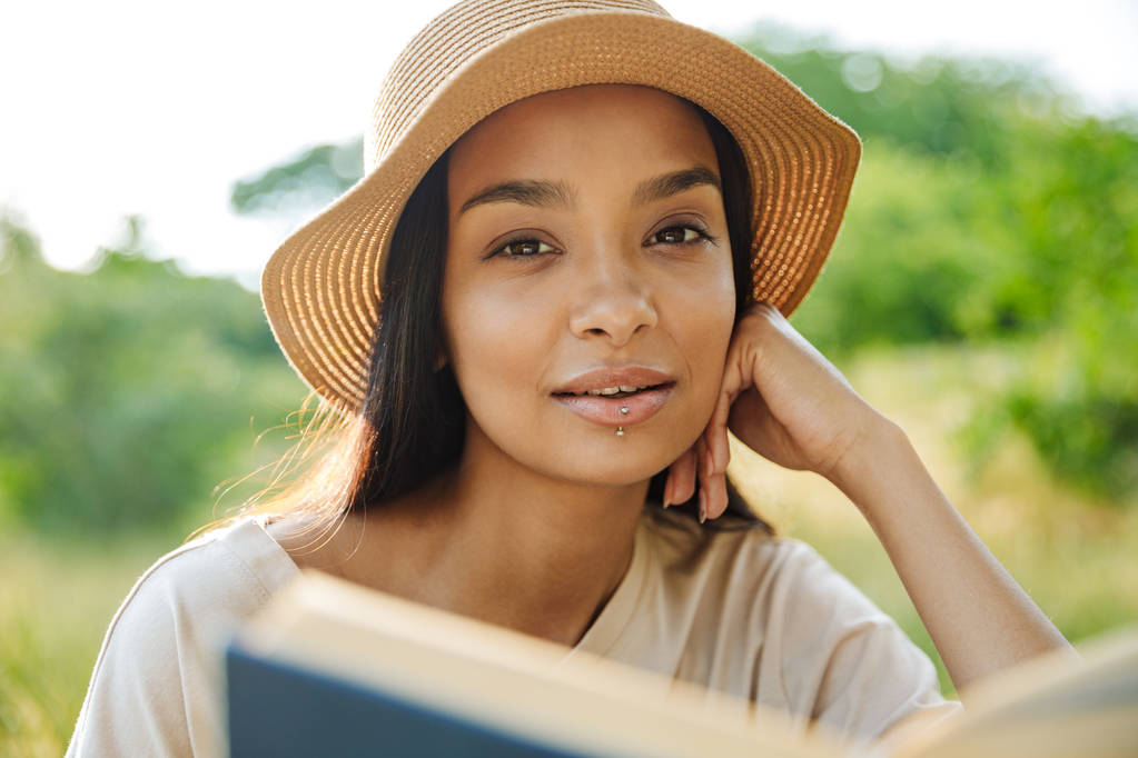 Muotokuva houkutteleva nainen yllään olki hattu käsittelyssä kirjan istuessaan ruohoa vihreässä puistossa
 - Valokuva, kuva