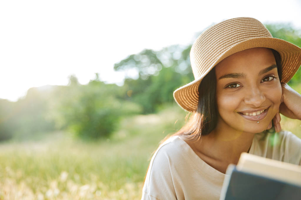 Portrait de femme joyeuse portant le livre de lecture de chapeau de paille tout en étant assis sur l'herbe dans le parc vert
 - Photo, image