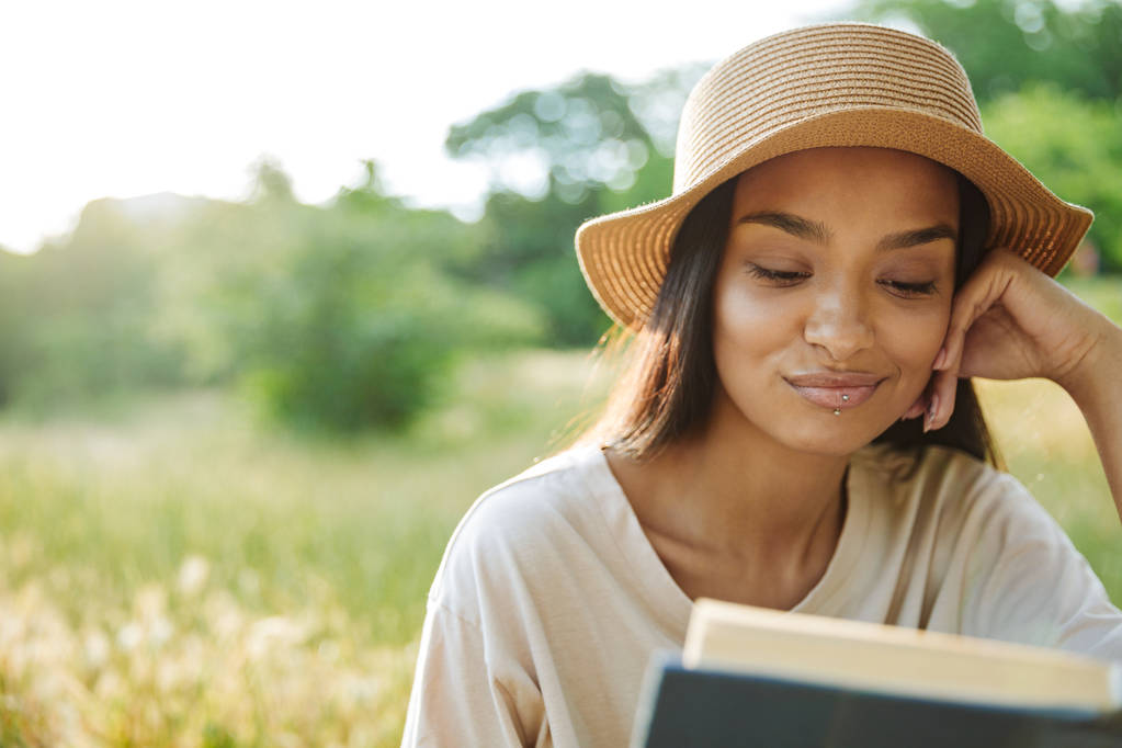 Портрет довольной женщины в соломенной шляпе, читающей книгу, сидящей на траве в зеленом парке
 - Фото, изображение