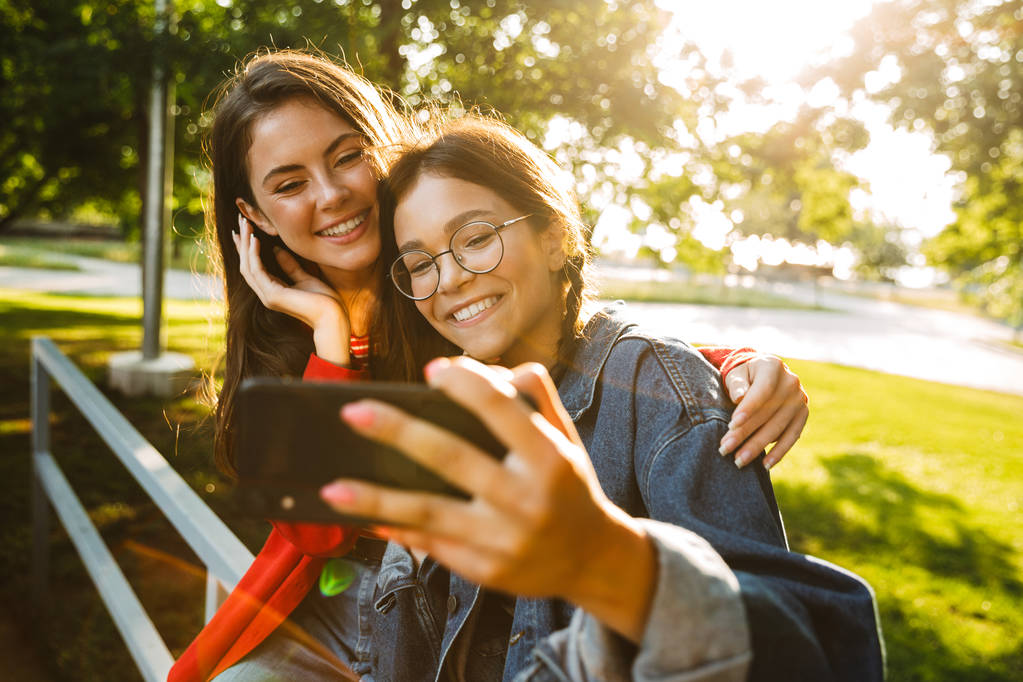 Imagen de dos chicas bonitas tomando una foto selfie en el teléfono celular y sonriendo mientras están sentadas en la barandilla en el parque verde
 - Foto, Imagen