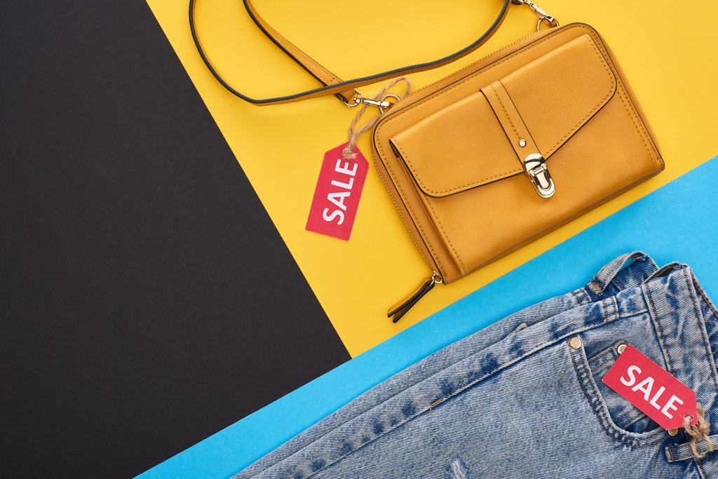 Draufsicht auf Tasche und Jeans mit Verkaufsetiketten auf blauem, gelbem und schwarzem Hintergrund - Foto, Bild