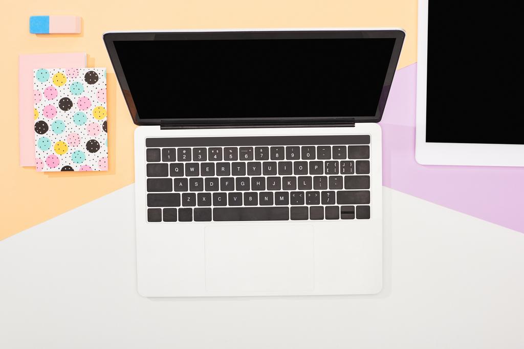 Draufsicht auf Laptop mit leerem Bildschirm, digitalem Tablet und Bürobedarf auf beigem, violettem und weißem Hintergrund - Foto, Bild