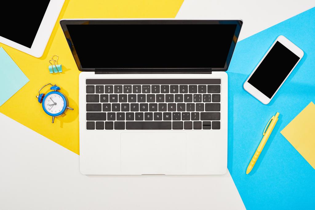 黄色、青、白の背景に事務用品を備えた空白の画面を持つラップトップ、スマートフォン、デジタルタブレットのトップビュー - 写真・画像