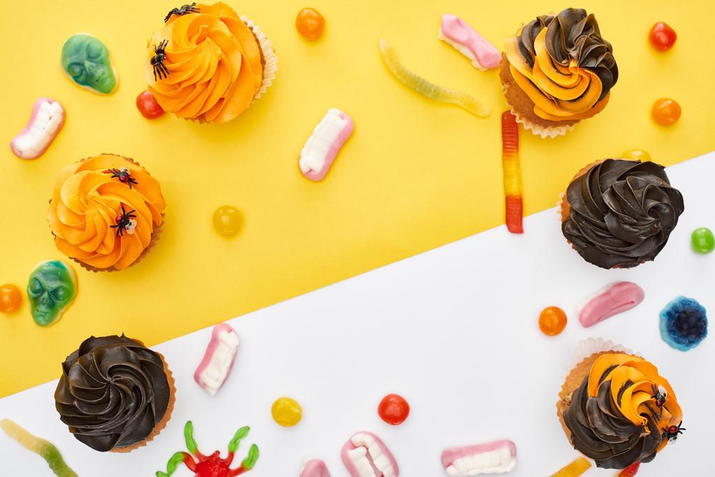 Draufsicht auf bunte Gummibärchen und Cupcakes auf gelb-weißem Hintergrund mit Kopierraum, Halloween-Leckerbissen - Foto, Bild
