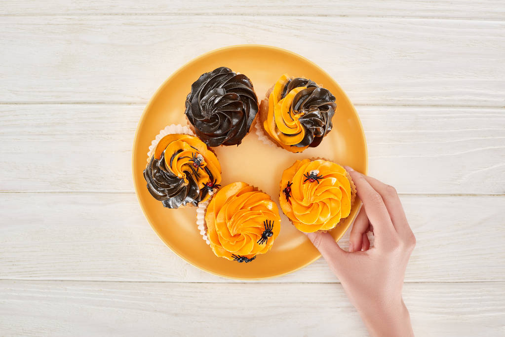 vista recortada de la mujer tomando delicioso cupcake con arañas de plato naranja en la mesa de madera blanca, regalo de Halloween
 - Foto, imagen