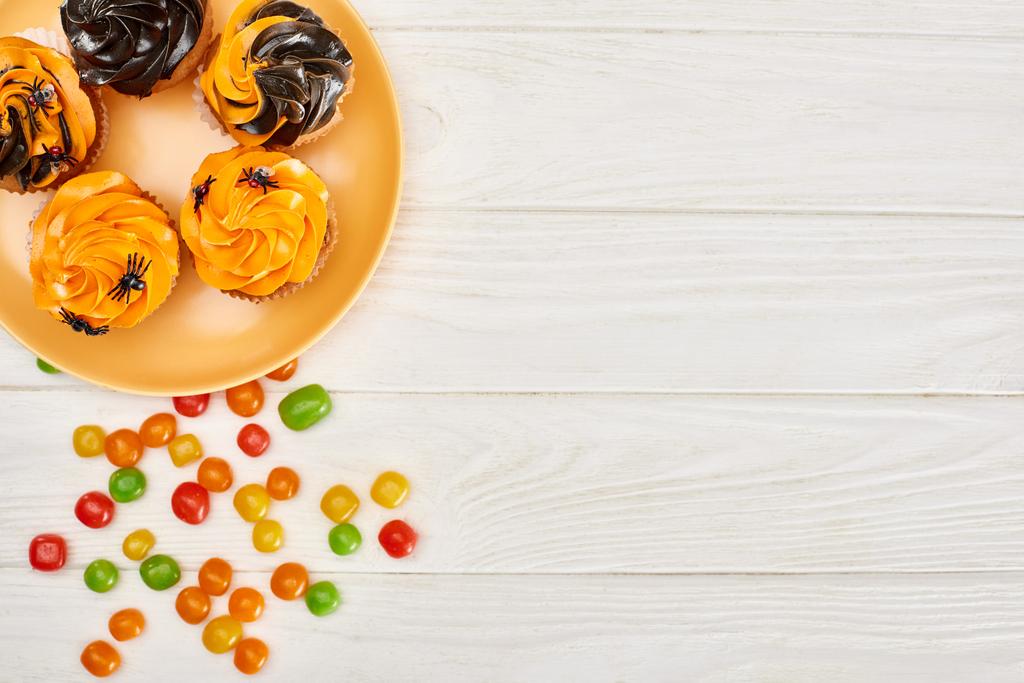 vista superior de bonbons coloridos perto da placa alaranjada com cupcakes na mesa de madeira branca, deleite de Halloween
 - Foto, Imagem