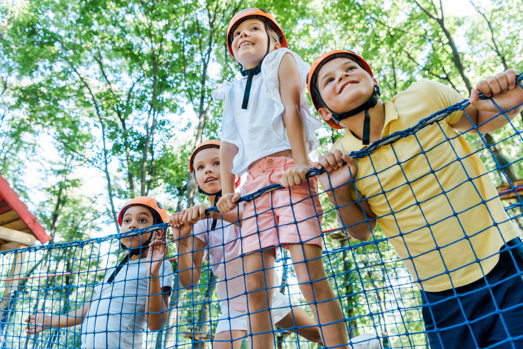 niski kąt widzenia uśmiechniętych dzieci w pomarańczowych hełmach w pobliżu wielokulturowych chłopców w parku przygody  - Zdjęcie, obraz