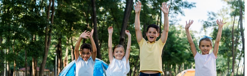 panoramiczne ujęcie szczęśliwych wielokulturowych dzieci z rękami nad głową  - Zdjęcie, obraz